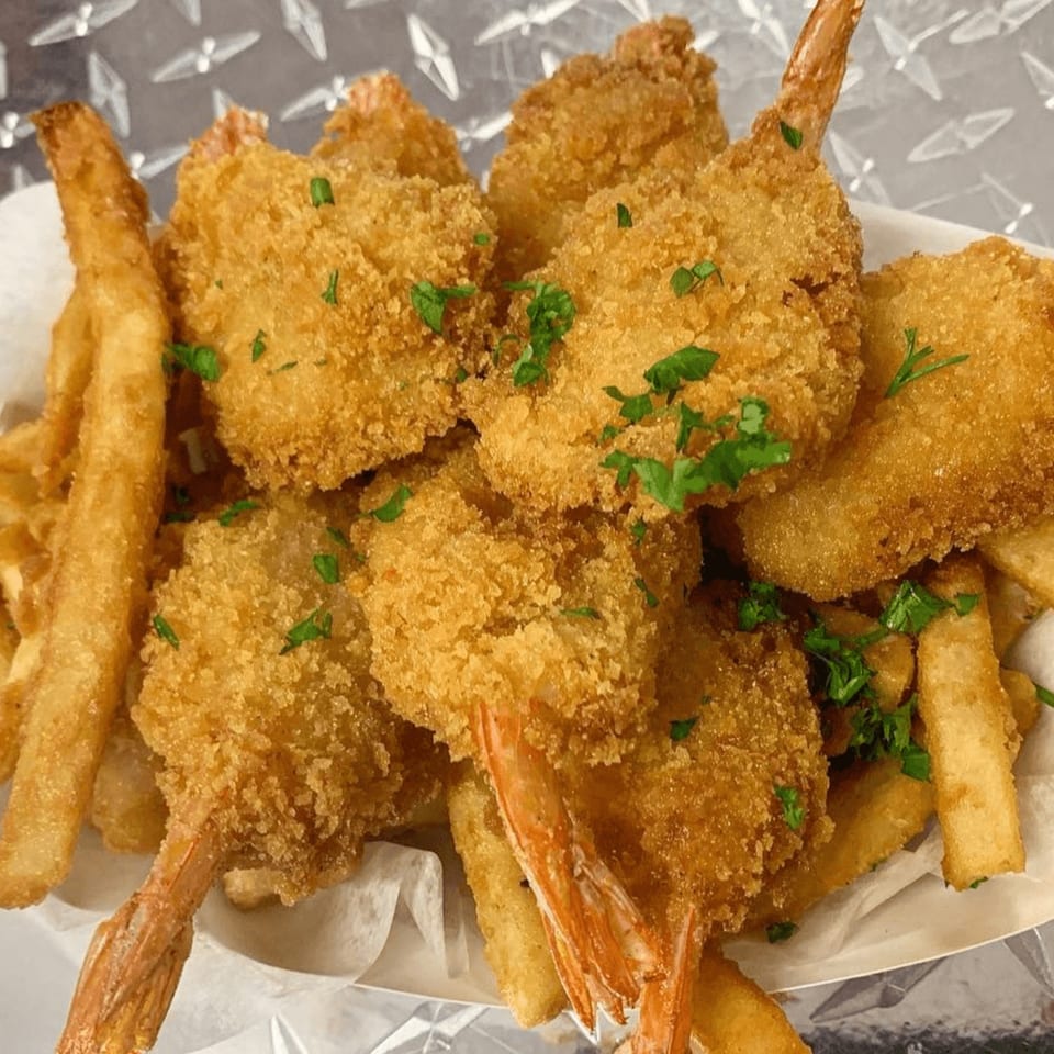 Golden-brown fried shrimp and french fries, garnished with parsley, served in a white paper tray.